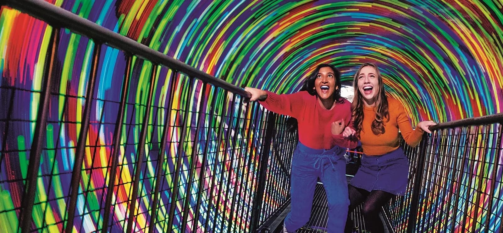 Two women laugh and grip the railings inside the colourful spinning Vortex Tunnel at Camera Obscura & World of Illusions in Edinburgh.