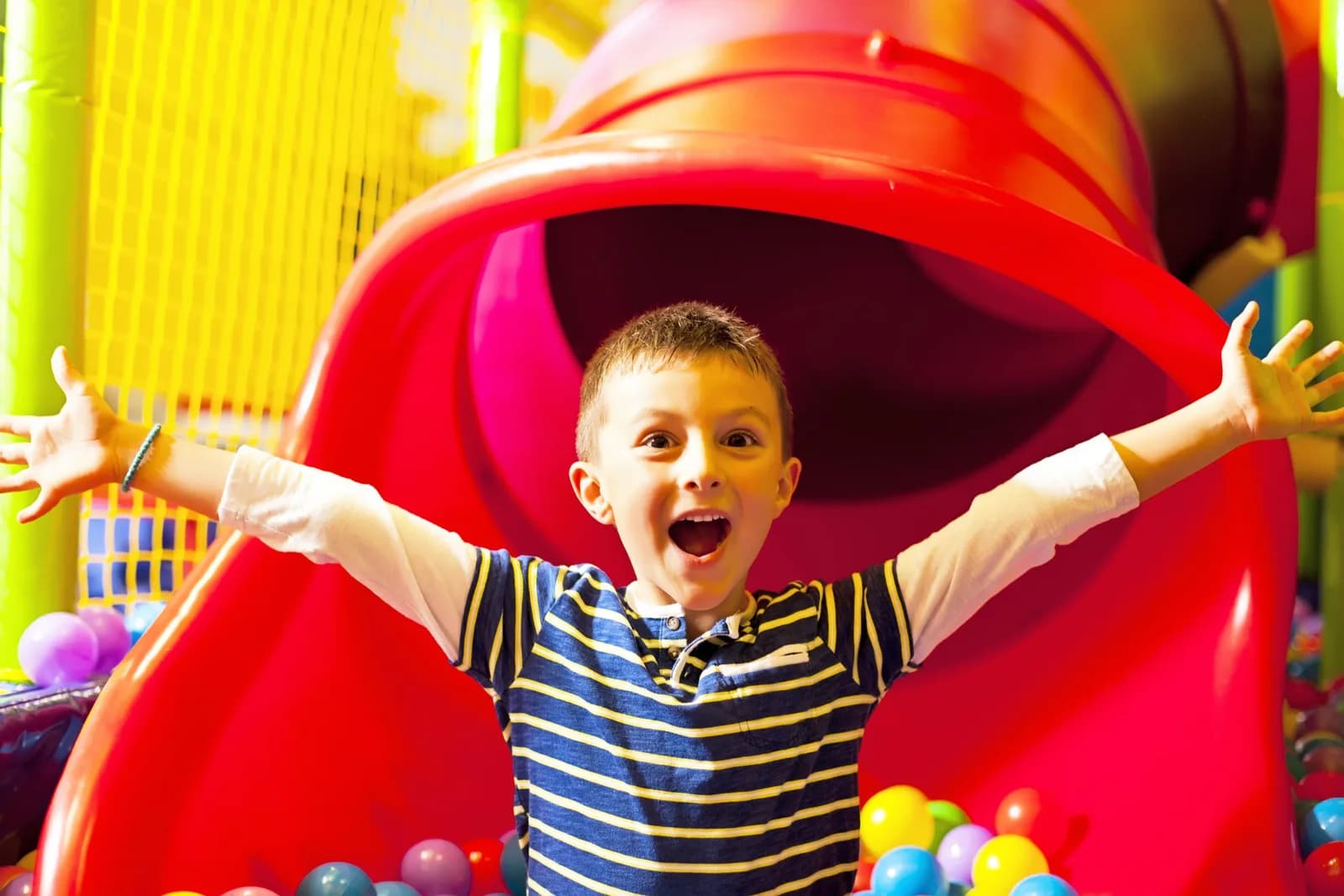 Child laughing at the base of a red tube slide in the Playzone Swansea soft play area, with ball pit and multi-level play frame behind