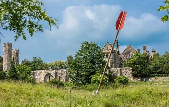 The giant arrow sculpture standing in the battlefield at 1066 Battle of Hastings, with the ruins of Battle Abbey and its gatehouse beyond